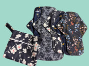 A set of reusable cloth pads and a matching zippered pouch laid out on a light fabric surface. The items feature dark navy backgrounds with white floral prints and multicolored floral motifs in pink, blue, yellow, and orange.